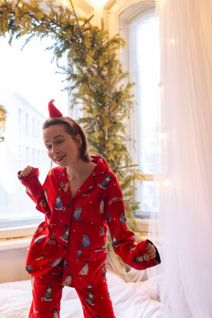 Cheerful funky young woman in nightwear jumping on bed on Christmas. Happy girl in red pajamas dancing alone at home in light roomの写真素材