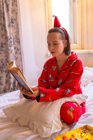 Pretty young woman reading book in bed on Christmas. Girl in red pajamas and Santa hat lying in bedroom. Cozy home atmosphereの写真素材