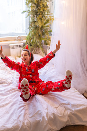 Happy girl in red pajamas lying alone in bed legs up at home in light bedroom. Christmas morning of young smiling womanの写真素材