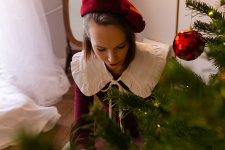 Portrait of beautiful girl decorating Christmas tree in living room. Woman dressed for Xmas in vintage burgundy dress with white colarand beret, view from aboveの写真素材