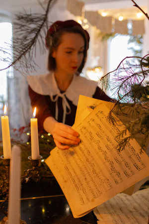Beautiful young woman in vintage burgundy beret and dress stands by piano decorated with Christmas branches and lights, reaching for sheet music. Cozy, festive and nostalgic holiday atmosphereの写真素材
