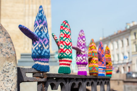Colorful handmade knitted mittens with traditional patterns on metal railing in city. Bright woolen gloves against urban architecture in background. Crafted accessories for autumn and winterの写真素材