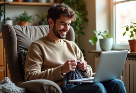 young man smiling while knitting near laptop, live host creating tutorial content and engaging audience; bright corner window, casual sweater, skein on lap, upbeat expression, modern home studio vibeの素材