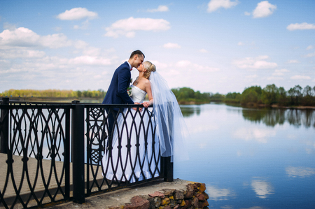 happy and beautiful groom and bride tender kiss at spring outdoors at the beach of the river under blue skyの写真素材