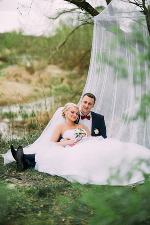 Elegant young happy wedding couple is sitting at green grass on green sunny summer background near the white canopy hugging, smiling and kissing.の写真素材