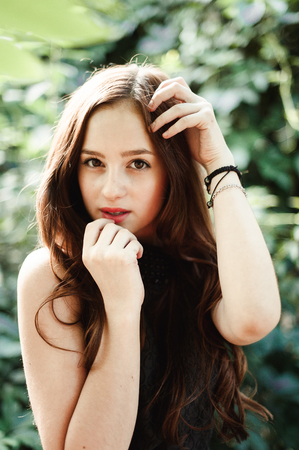 Beautiful young girl in black dress with long hair, super cute and natural standing among colorful green leavesの写真素材