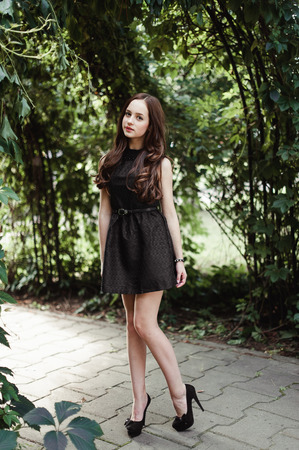 Beautiful young girl in black dress with long hair, super cute and natural standing among colorful green leavesの写真素材
