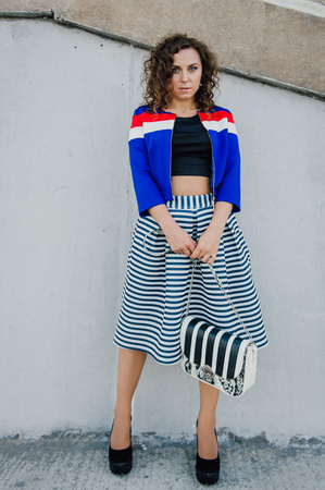 Beautiful sexy young brunette girl with long wavy hair thin slender figure and pretty face make-up wearing a long stripped skirt and black top, high heels. Walk on the street in casual styleの写真素材