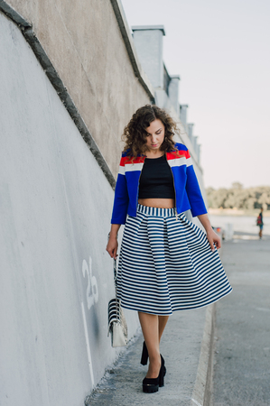 Beautiful sexy young brunette girl with long wavy hair thin slender figure and pretty face make-up wearing a long stripped skirt and black top, high heels. Walk on the street in casual styleの写真素材