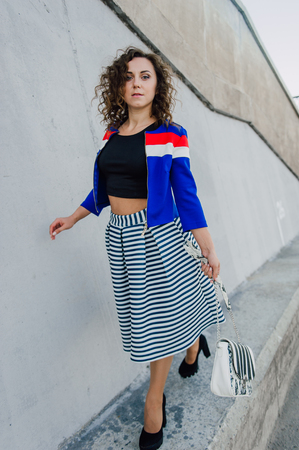 Beautiful sexy young brunette girl with long wavy hair thin slender figure and pretty face make-up wearing a long stripped skirt and black top, high heels. Walk on the street in casual styleの写真素材