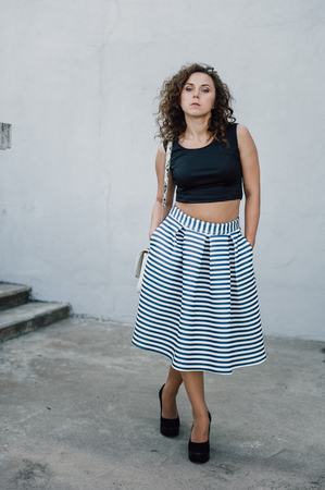 Beautiful sexy young brunette girl with long wavy hair thin slender figure and pretty face make-up wearing a long stripped skirt and black top, high heels. Walk on the street in casual styleの写真素材