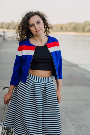 Beautiful sexy young brunette girl with long wavy hair thin slender figure and pretty face make-up wearing a long stripped skirt and black top, high heels. Walk on the street in casual styleの写真素材