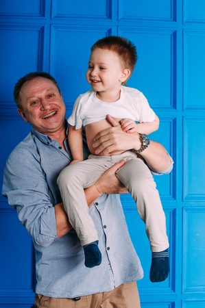Laughing grandfather with his grandson as they play together indoors in the living room with the cute young boy hugging him from behindの写真素材