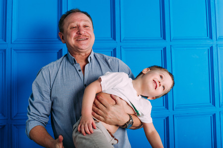 Laughing grandfather with his grandson as they play together indoors in the living room with the cute young boy hugging him from behindの写真素材