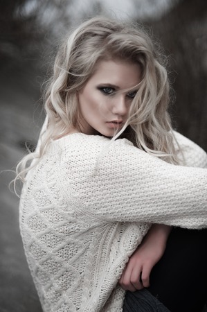 Close up of beautiful young  blonde woman with blue eyes in white pullover, sitting.
Winter, spring, outdoor portrait. Professional beauty make-up: dark smoky eyes and pale lips and hair style.の写真素材