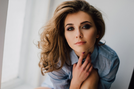 Young pretty woman sitting at window and looking outside enjoys of rest, wearing blue shirt. Studio retouched shot.の写真素材