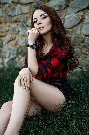 Beautiful young hipster girl posing and smiling near urban wall background in red plaid shirt, shorts, outdoors summer portrait.の写真素材