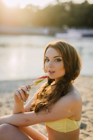 summer vacation, travel, tourism, junk food and people concept - young woman in sunglasses eating colorful ice cream on resort beach background, wearing yellow swimsuitの写真素材