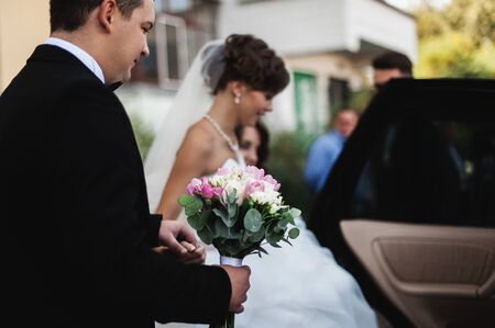 Elegant young happy wedding couple is sitting at green grass on green sunny summer background near the white canopy hugging, smiling and kissing.の写真素材
