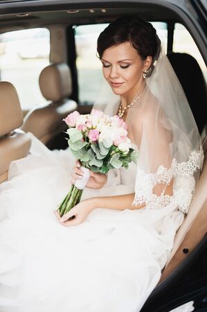 Elegant young happy wedding couple is sitting at green grass on green sunny summer background near the white canopy hugging, smiling and kissing.の写真素材