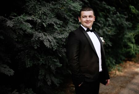 Elegant young happy wedding couple is sitting at green grass on green sunny summer background near the white canopy hugging, smiling and kissing.の写真素材
