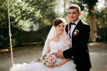Elegant young happy wedding couple is sitting at green grass on green sunny summer background near the white canopy hugging, smiling and kissing.の写真素材
