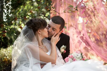 Elegant young happy wedding couple is sitting at green grass on green sunny summer background near the white canopy hugging, smiling and kissing.の写真素材