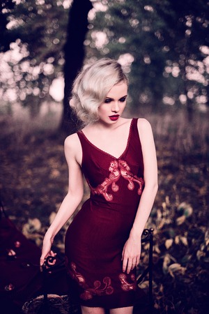 Beautiful and elegant blonde woman with red lips and hair waves wearing wine red nightie posing on the bed outdoors, retro vintage style and fashionの写真素材