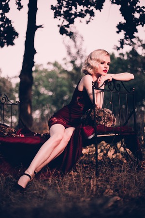 Beautiful and elegant blonde woman with red lips and hair waves wearing wine red nightie posing on the bed outdoors, retro vintage style and fashionの写真素材