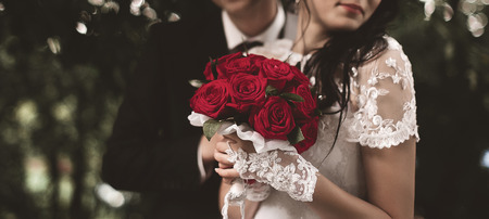 Elegant young happy wedding couple is sitting at green grass on green sunny summer background near the white canopy hugging, smiling and kissing.の写真素材