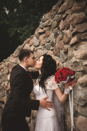 Elegant young happy wedding couple is sitting at green grass on green sunny summer background near the white canopy hugging, smiling and kissing.の写真素材
