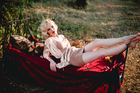 Beautiful and elegant blonde woman with red lips and hair waves wearing wine red nightie posing on the bed outdoors, retro vintage style and fashionの写真素材