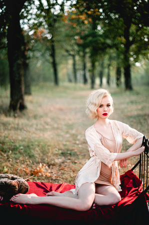 Beautiful and elegant blonde woman with red lips and hair waves wearing wine red nightie posing on the bed outdoors, retro vintage style and fashionの写真素材
