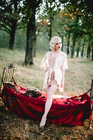 Beautiful and elegant blonde woman with red lips and hair waves wearing wine red nightie posing on the bed outdoors, retro vintage style and fashionの写真素材