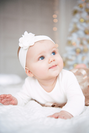 Baby girl wearing cute dress and headband, lies on a white cover in festively decorated room. With surprise watches in the camera, on a background a set of bright fires, soft focus. Warm beige and gold colorsの写真素材