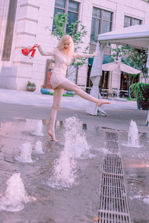 Beautiful young blond woman with hair in a dress near a fountain in a summer dayの写真素材