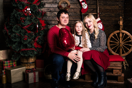 Beautiful young family in red having fun together for Christmas holidays, sitting on a living room floor next to a nicely decorated Christmas tree, smilingの写真素材