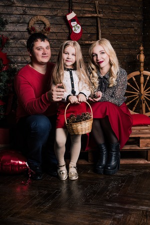 Beautiful young family in red having fun together for Christmas holidays, sitting on a living room floor next to a nicely decorated Christmas tree, smilingの写真素材