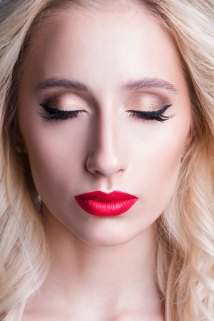 Fashion beautiful woman with blond curly hair. Beautiful portrait of sensual young woman model with glamour red lips make-up, closed eyes with arrow makeup, purity skinの写真素材