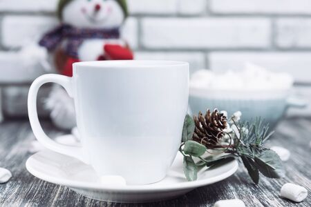 Drink in a white ceramic cup with zephyr on wooden table. Decorative pine branch with the cone on plate, saucer. Snowman on backgroundの写真素材
