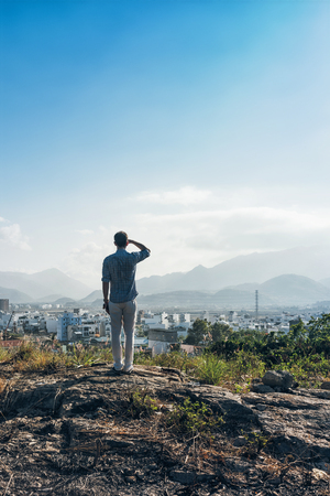 Man on a top of a hill. Fashionable man in white trousers and blue shirt enjoying city and mountains viewの写真素材