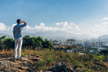 Man on a top of a hill. Fashionable man in white trousers and blue shirt enjoying city and mountains viewの写真素材