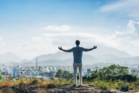 Man on a top of a hill. Fashionable man in white trousers and blue shirt standing with raised hands and enjoying city and mountains viewの写真素材