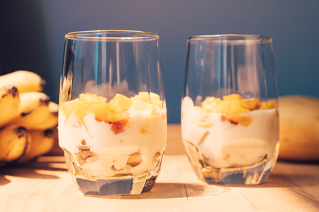 Glasses of yoghurt with mango and strawberry. Homemade healthy yogurt with fruits. A banch of bananas on a table. Morning sunlightの写真素材