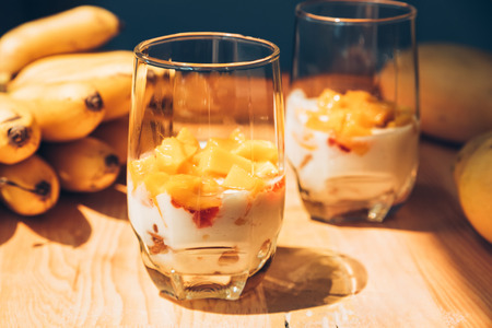 Glasses of yoghurt with mango and strawberry. Homemade healthy yogurt with fruits. A banch of bananas on a table. Morning sunlightの写真素材