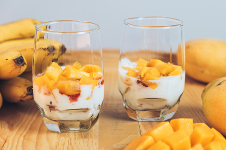 Glasses of yoghurt with mango and strawberry. Homemade healthy yogurt with fruits. A banch of bananas on a table. Morning sunlightの写真素材