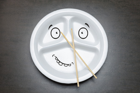 Empty disposable plastic plate on black table. White plate with three sections for food with spoon and bamboo chopsticks. Happy smiley drawn face on dish plateの写真素材
