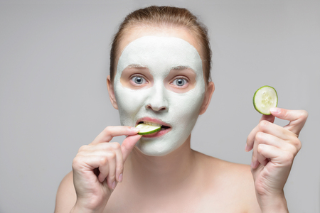 Girl with green cream on face. Beautiful young woman with homemade mask biting a cucumberの写真素材