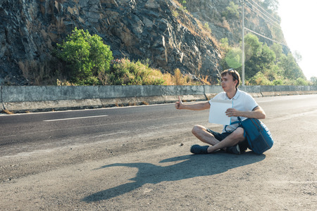 Young man hitchhiker sitting on the road. Tired man with blue backpack holding a white blank cardboard sign. Hitchhiking at sunset or sunriseの写真素材