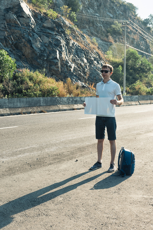 Young man hitchhiker standing on the road. Happy man with blue backpack holding a white blank cardboard sign. Hitchhiking at sunset or sunriseの写真素材
